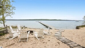 Een strand met witte Adirondack-stoelen in een halve cirkel, een vuurkuil en een houten looppad dat naar een steiger leidt.