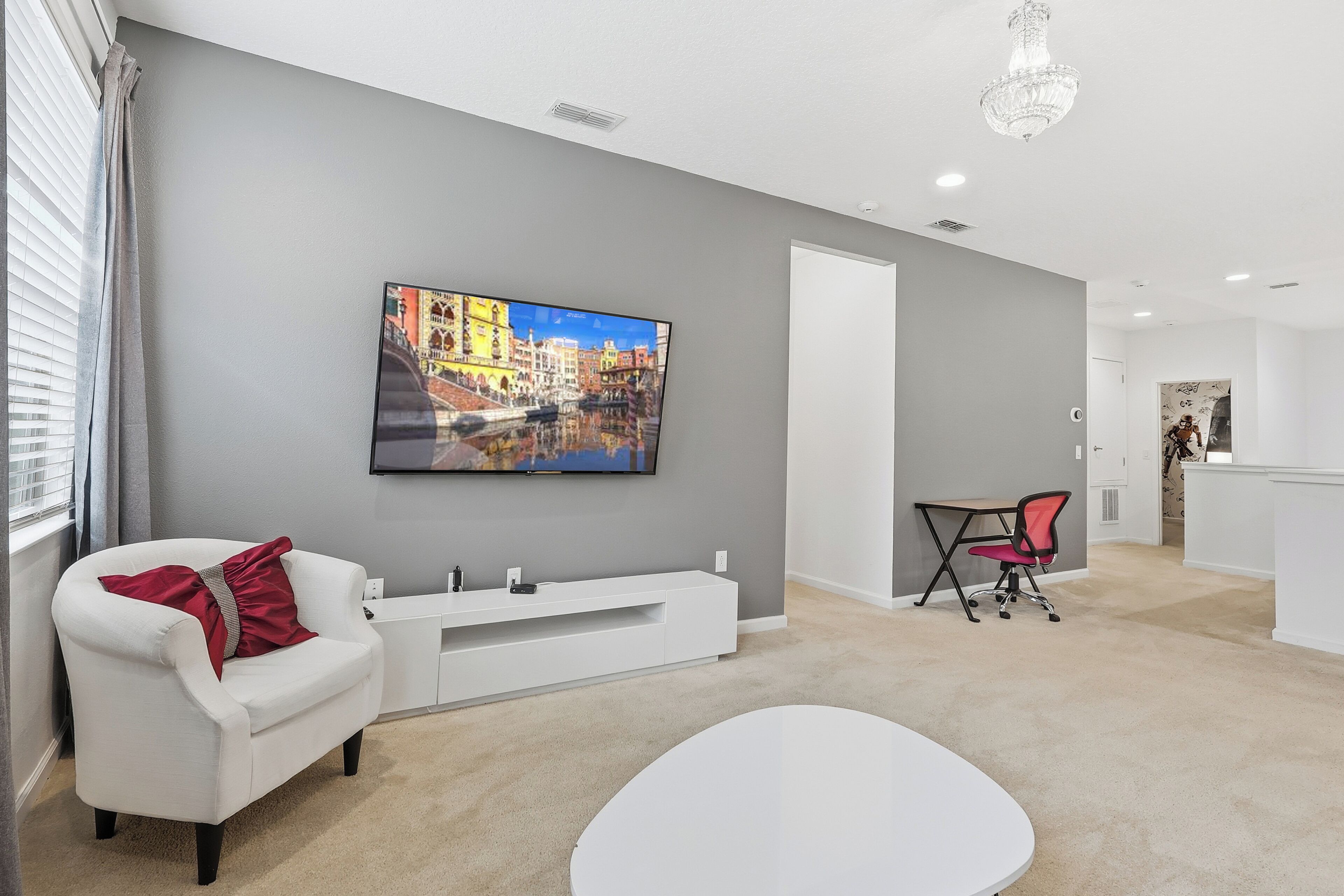 A modern living room with a flat-screen TV mounted on the wall, a white sofa with a red pillow, a small white table, and a red office chair.
