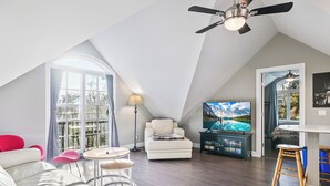 Un loft spacieux avec un plafond haut, un ventilateur de plafond, une grande fenêtre avec des rideaux, un canapé blanc, une petite table ronde, un téléviseur et un tabouret en bois.