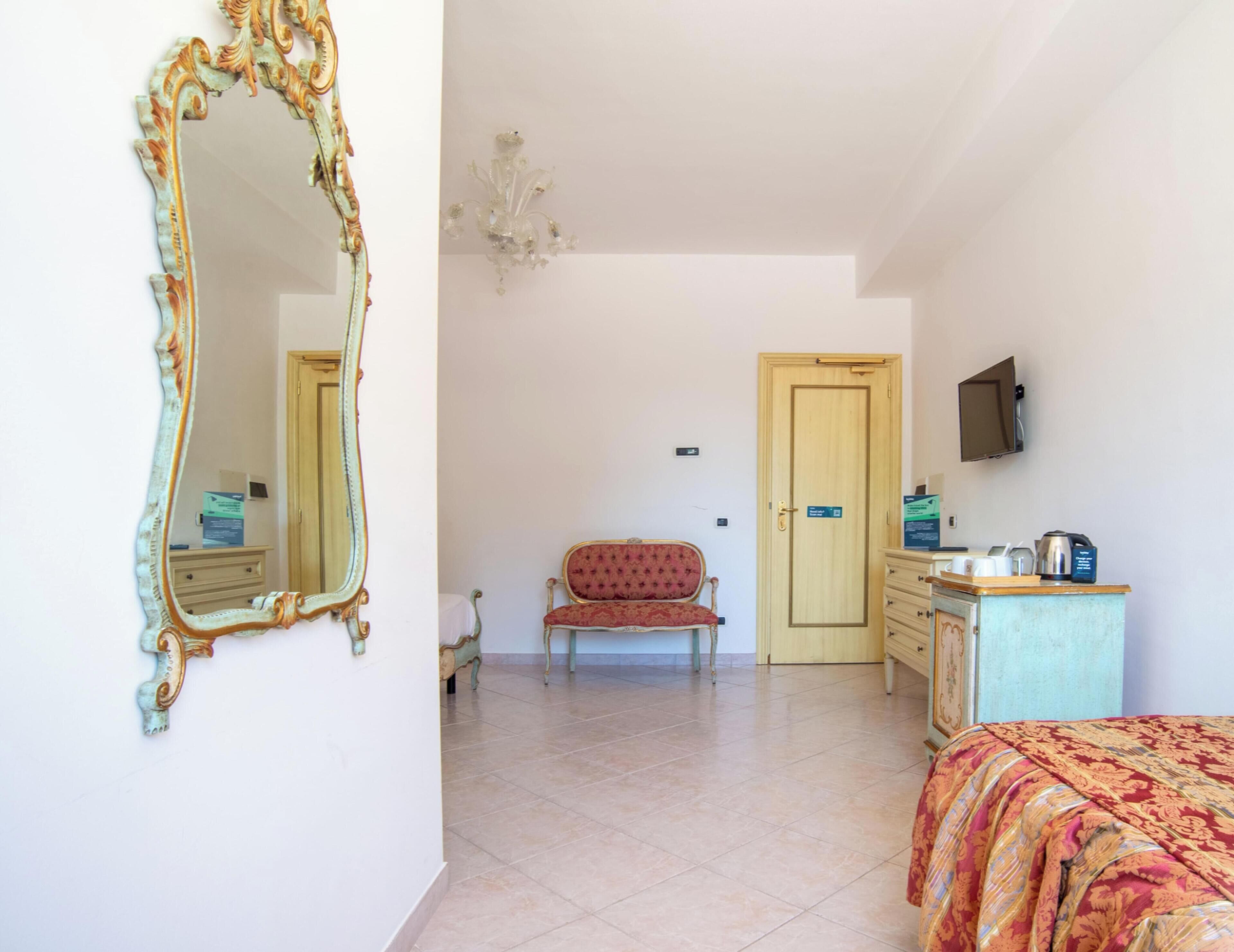 Um quarto com um espelho grande e ricamente decorado, uma poltrona estofada vermelha, uma porta de madeira, uma televisão fixada na parede e uma cama com colcha estampada.
