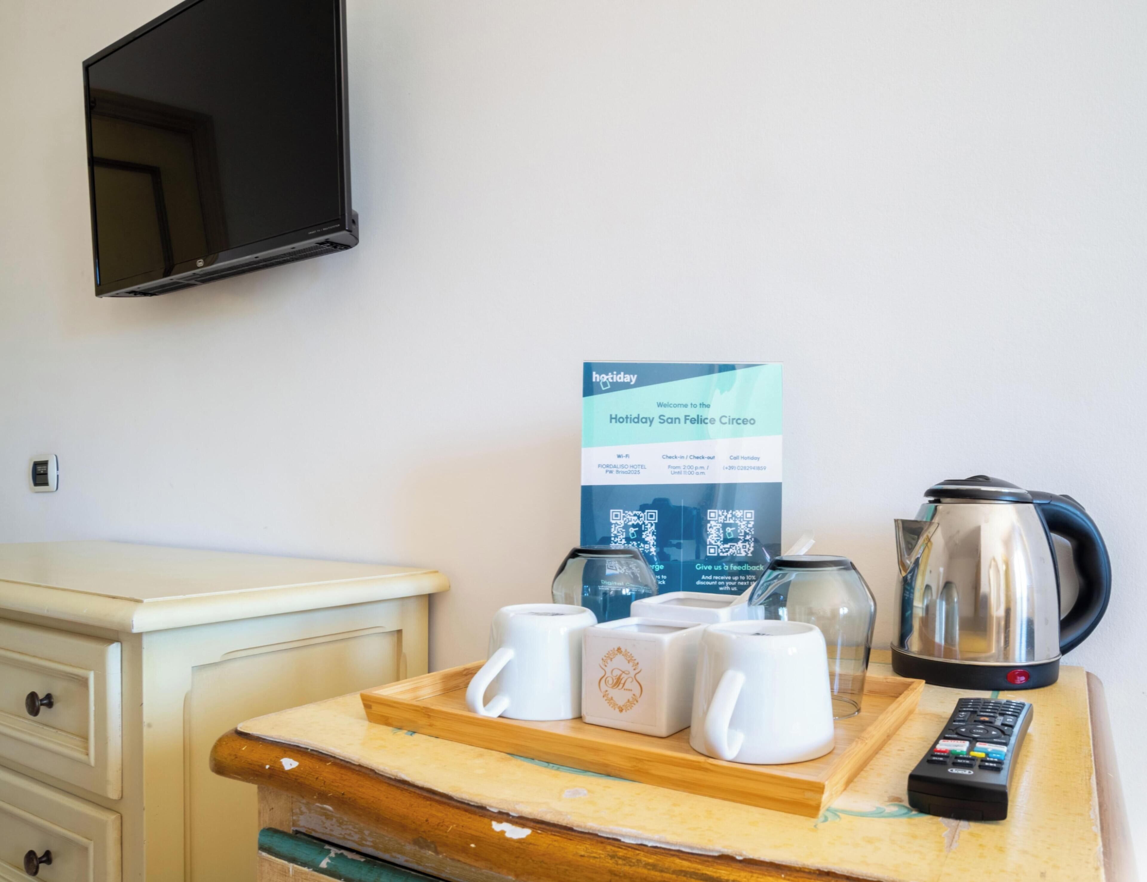 A hotel room with a wooden tray holding white mugs, a kettle, and a remote control on a wooden table.