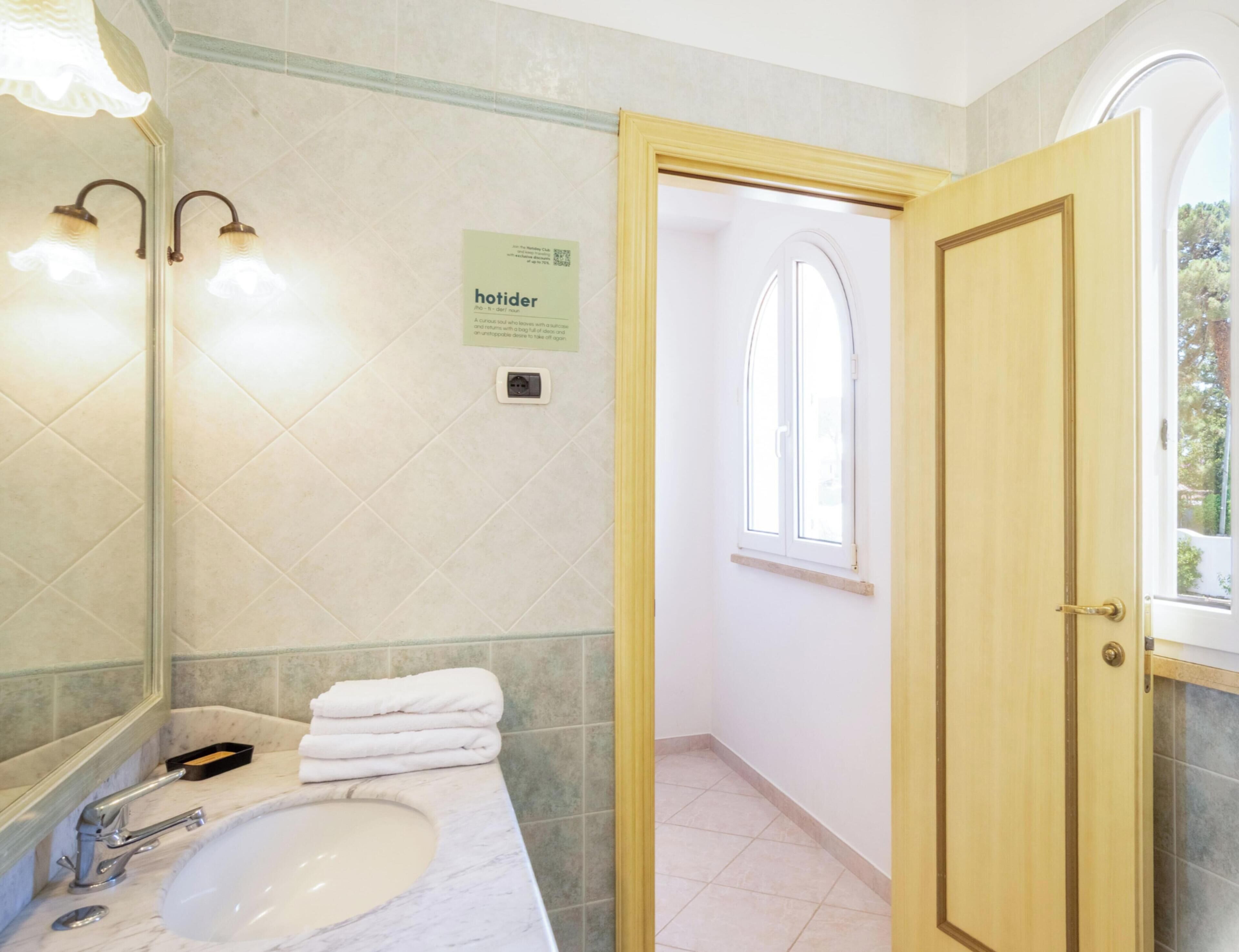 Un bagno con lavandino, specchio, porta asciugamani e un cartello appeso al muro.