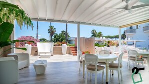 Une terrasse extérieure couverte, dotée de meubles blancs, d’un ventilateur de plafond et offrant une vue sur la mer et les bâtiments.