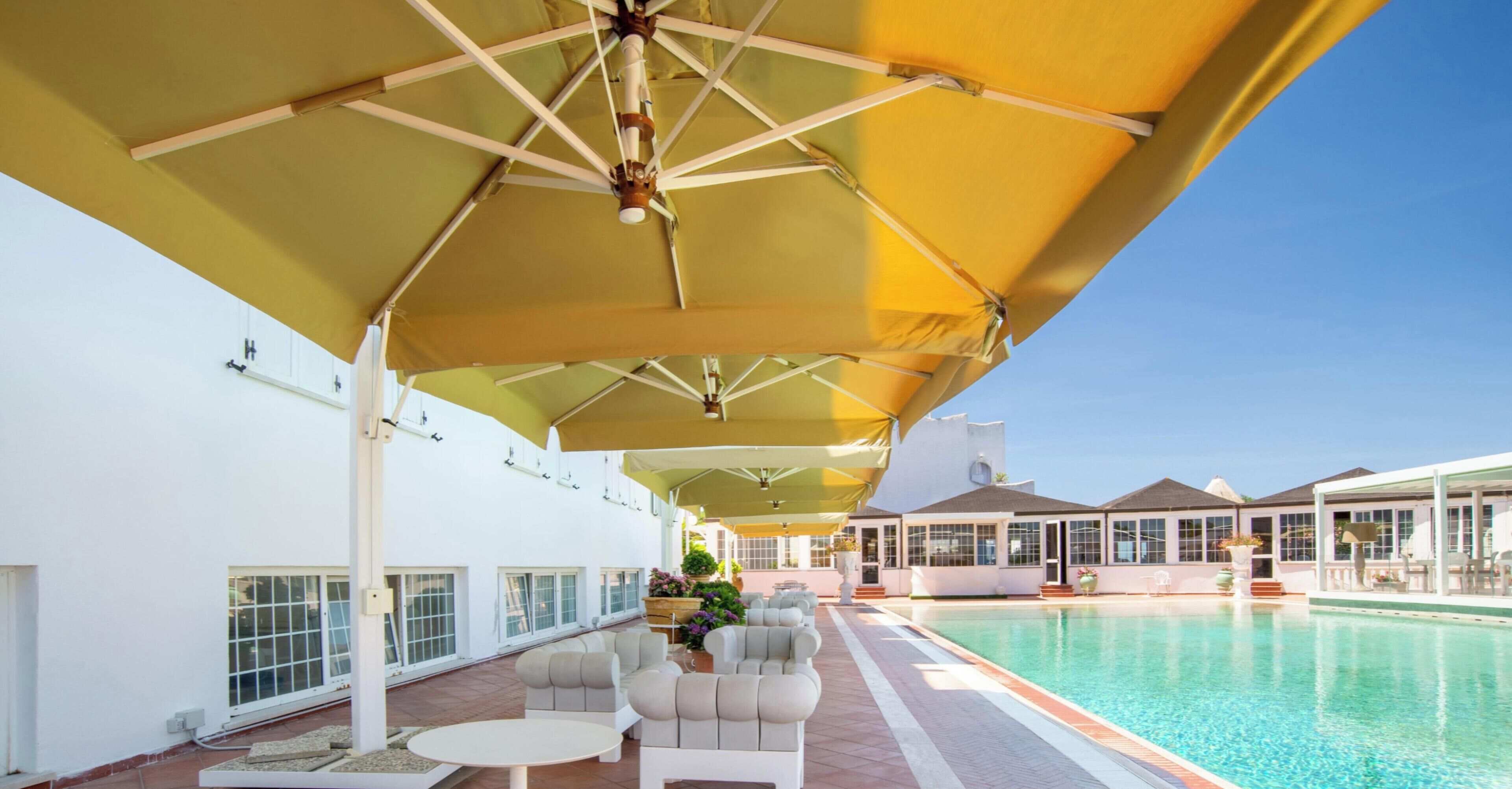 Un espace aménagé au bord de la piscine, avec des sièges et des parasols, une piscine et un bâtiment doté de grandes fenêtres.
