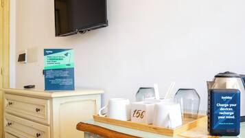 A hotel room with a TV mounted on the wall, a minibar, a tray with glasses and a charger, and a sign promoting travel ideas.