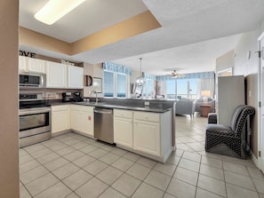 A modern kitchen with white cabinets, a built-in oven, and a dishwasher. There is a dining area with a gray sofa and a small table. - Gulf Front 3/3 Summerwind 9th floor. Sunny and Chair (Navarre Beach)