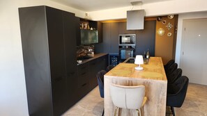 A modern kitchen with dark cabinetry, a central island, and a dining area with a wooden table and chairs. - L'Eden Palavas 70m2* Sublime T3*Parking*Terrace*Beach (Palavas-les-Flots)