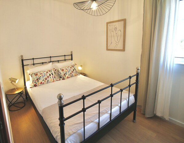 A neatly made bed with a black metal frame, a round bedside table with a lamp, and a framed artwork on the wall. - Desertas Beach and Sport (Ilhavo)