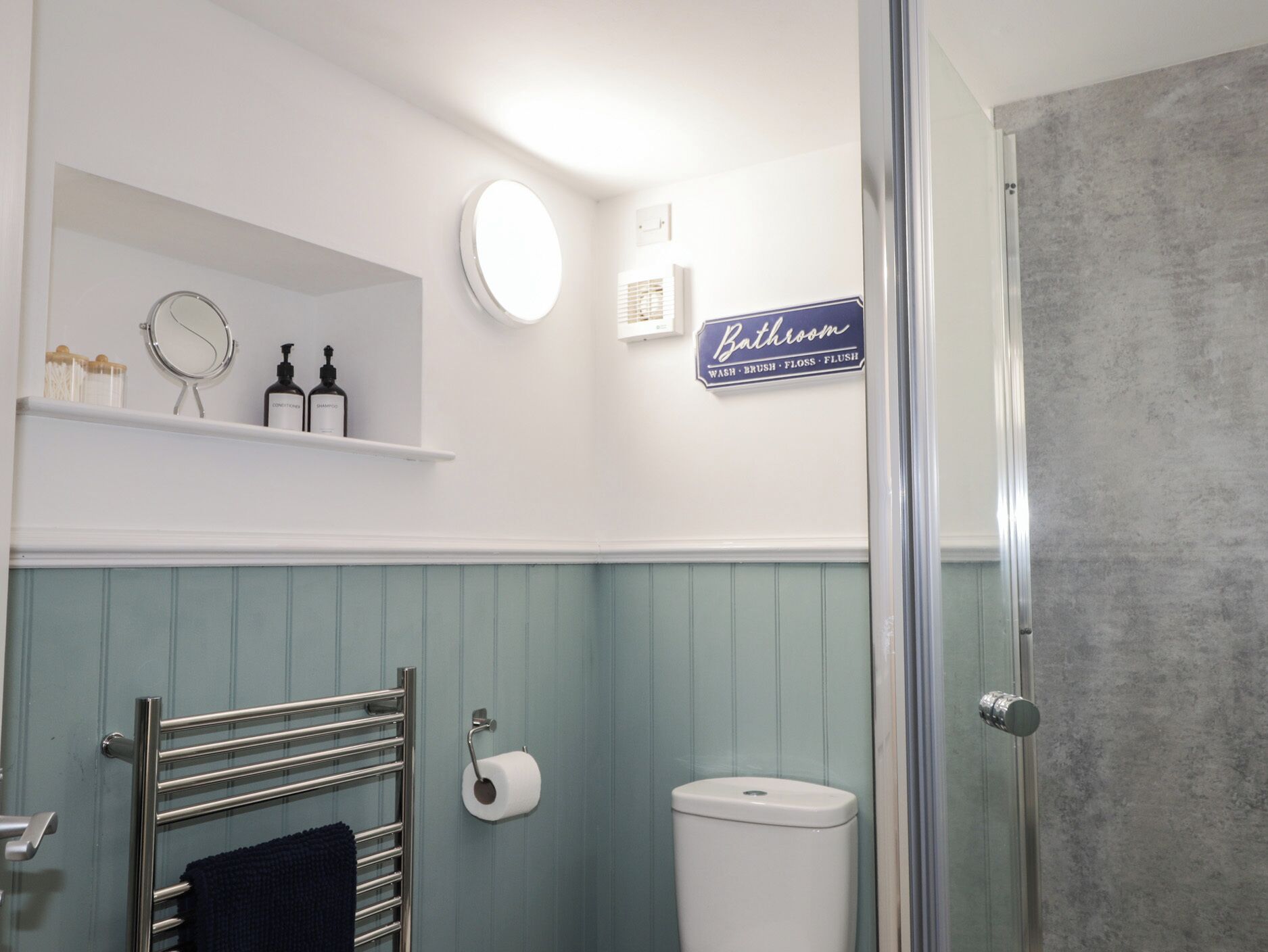 Un baño con inodoro, perchero para toallas, repisa con artículos de aseo, espejo y señalización que indica las funciones del baño.
