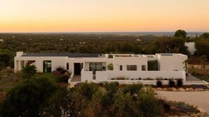 Um edifício moderno branco, com telhado plano, rodeado por vegetação e céu limpo.