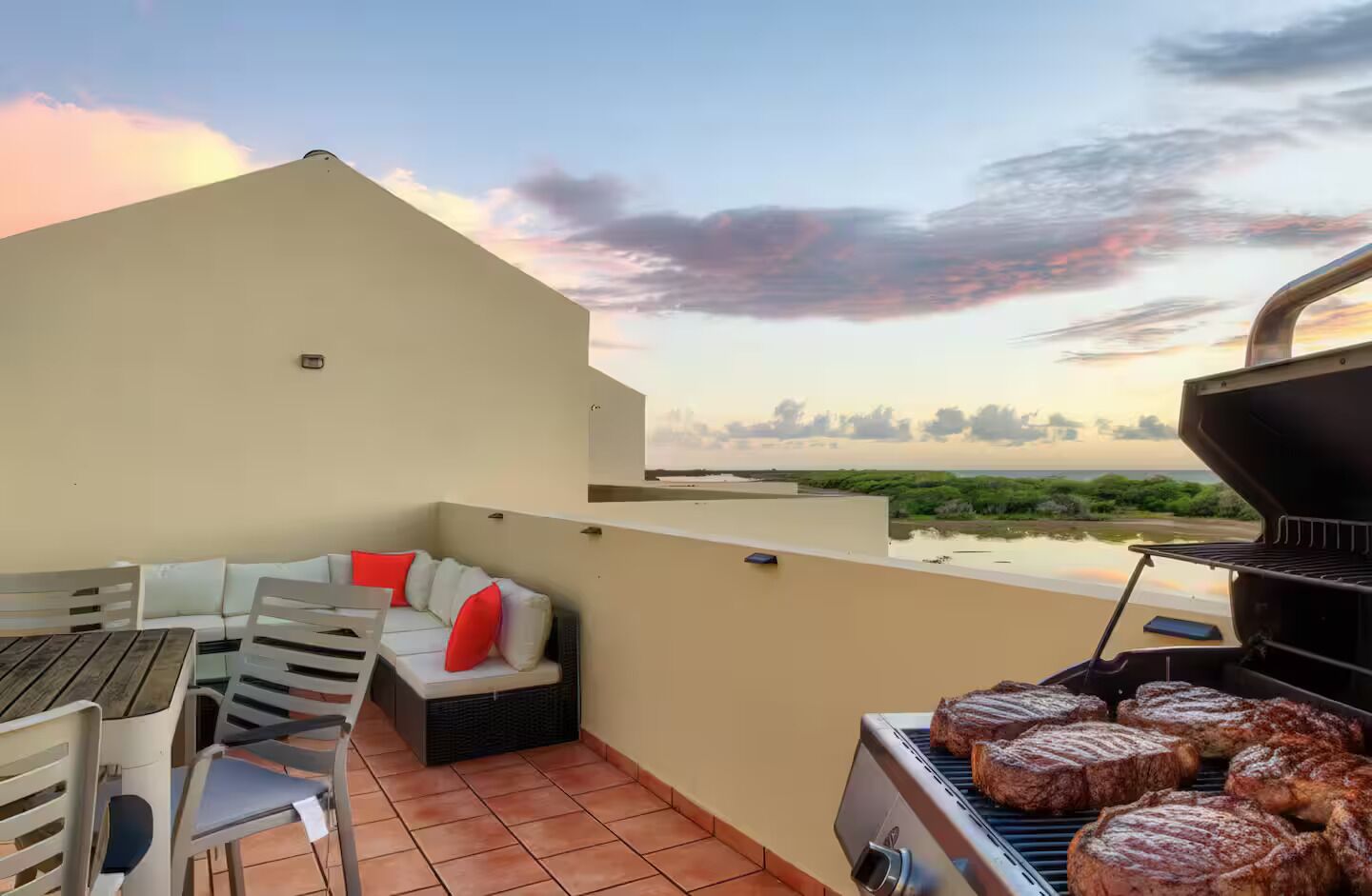 Una terraza en la azotea con una parrilla, mobiliario exterior y vistas a la playa al atardecer.