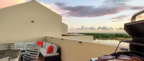 Una terraza en la azotea con una parrilla, mobiliario exterior y vistas a la playa al atardecer.