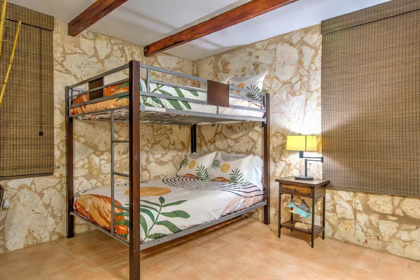 A bunk bed room with a stone wall, a small table with a lamp, and a window with a blind.