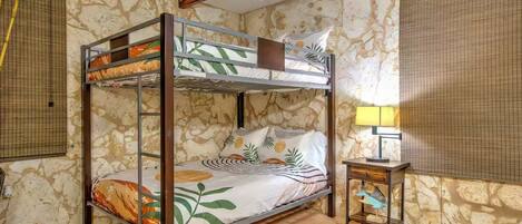 A bunk bed room with a stone wall, a small table with a lamp, and a window with a blind.