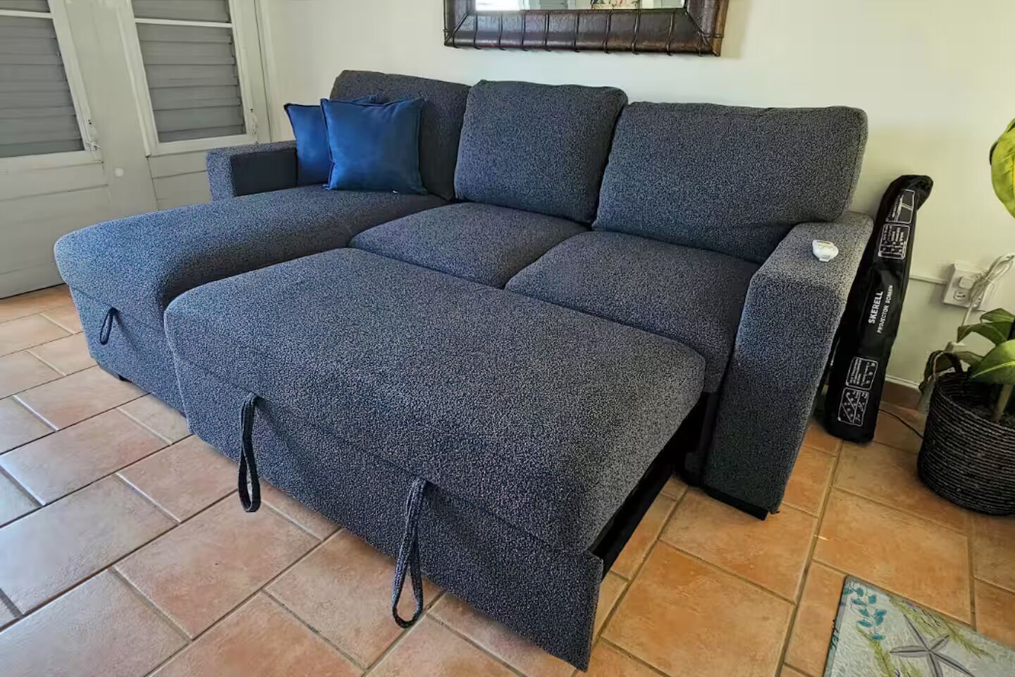 Un canapé gris avec un repose-pieds, un sac noir et une plante en pot dans un coin.
