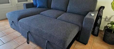 Un canapé gris avec un repose-pieds, un sac noir et une plante en pot dans un coin.