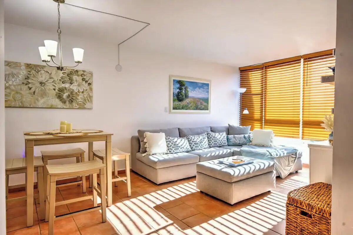 A living room with a sofa, ottoman, and a dining table with chairs. There is a painting on the wall and a window with blinds.
