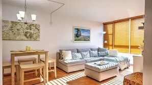 Ein Wohnzimmer mit einer Couch, einem Hocker und einem Esstisch mit Stühlen. An der Wand hängt ein Gemälde und es gibt ein Fenster mit Jalousien.