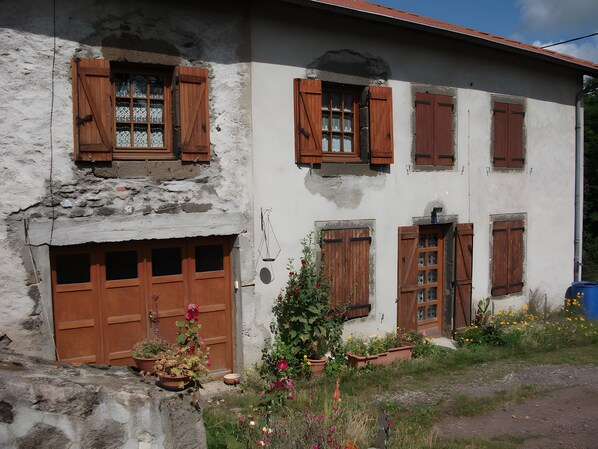A rustic building with wooden shutters, a stone wall, and a garden with potted plants.