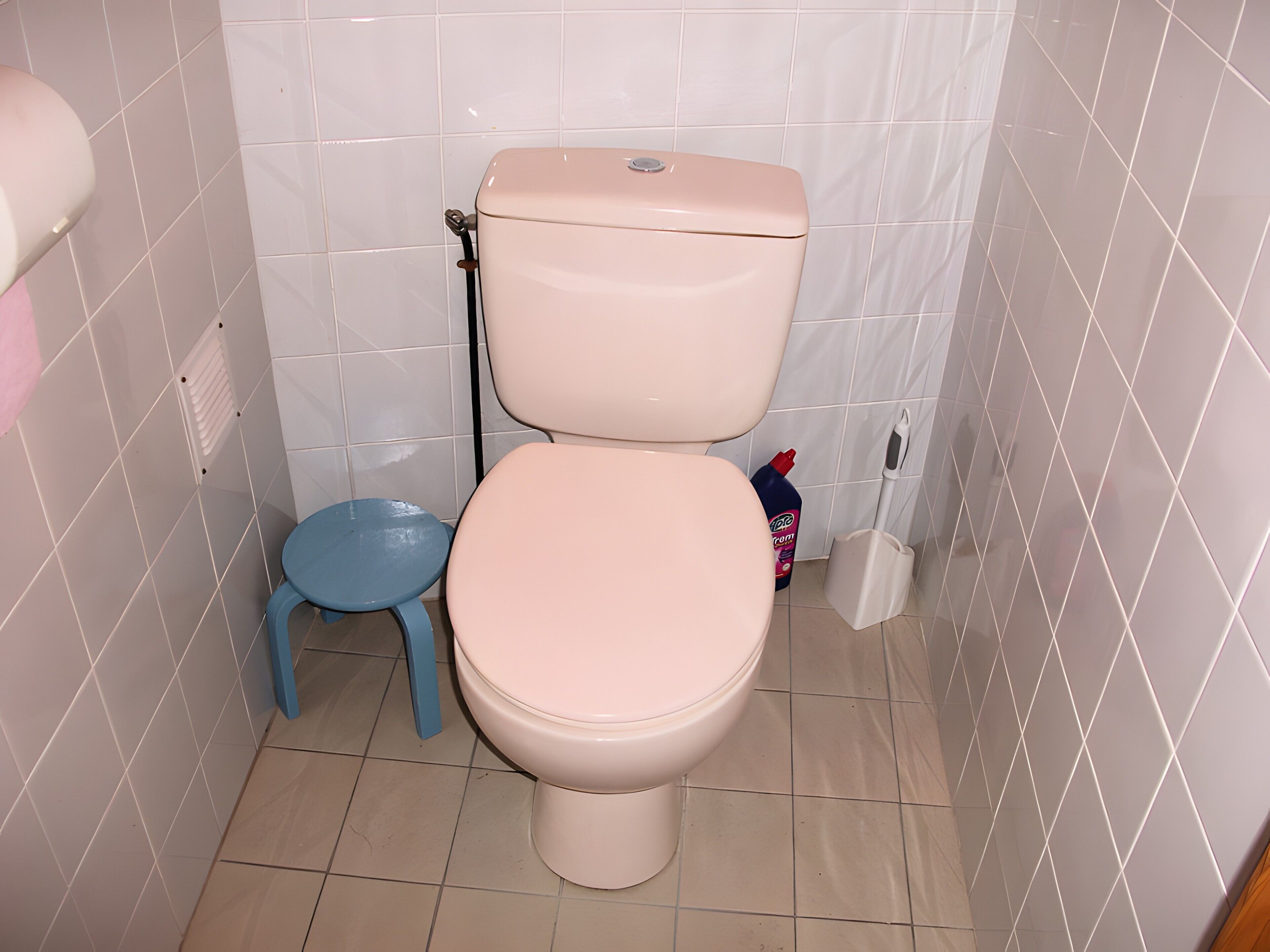 A toilet with a tank and seat, a blue plastic stool, and a bottle of cleaning product.