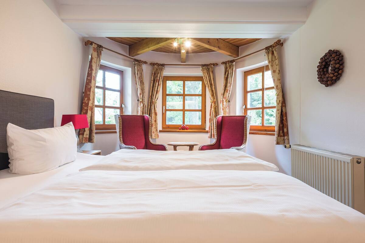 A large bed with white linens, two red upholstered chairs, a small table, and a window with curtains.