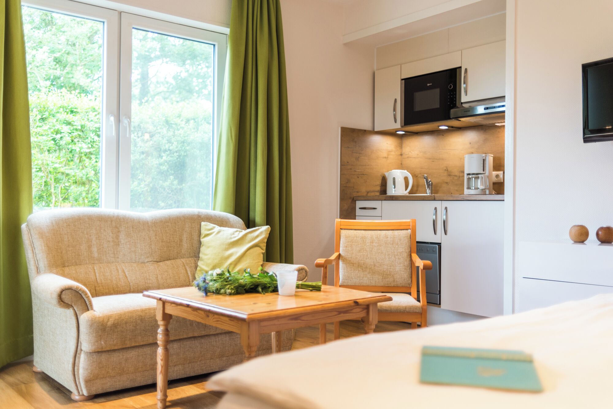 A cozy room with a sofa, a small wooden table, a chair, a bed, and a kitchenette with a microwave and coffee maker.