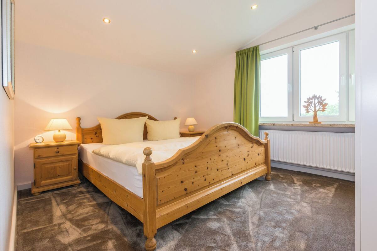 A bedroom with a wooden bed, two bedside tables with lamps, a window with green curtains, and a radiator.
