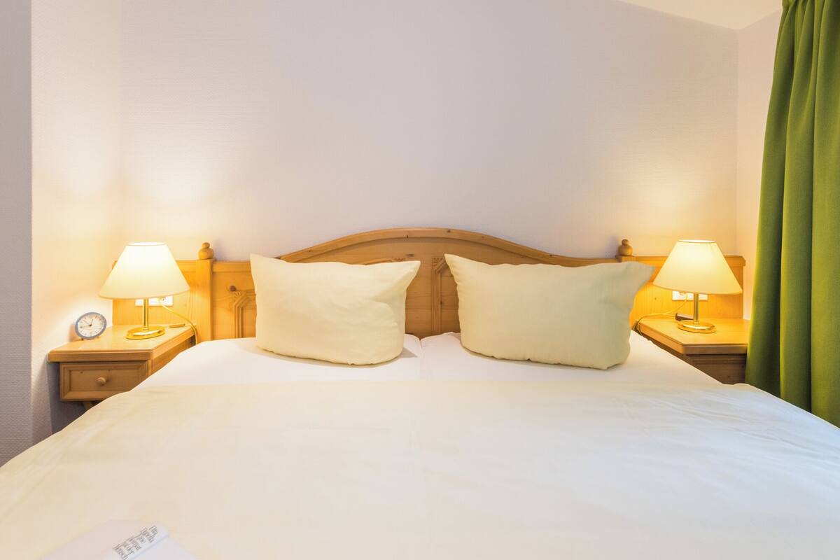 A neatly made bed with two pillows, a wooden headboard, and two bedside lamps on wooden nightstands.