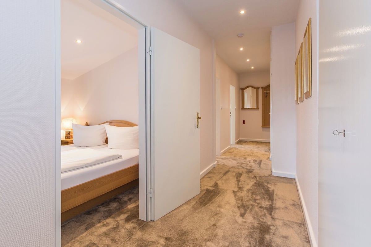 A hotel room with a bed, a nightstand, and a lamp. There is a hallway leading to another room with a mirror and a door.