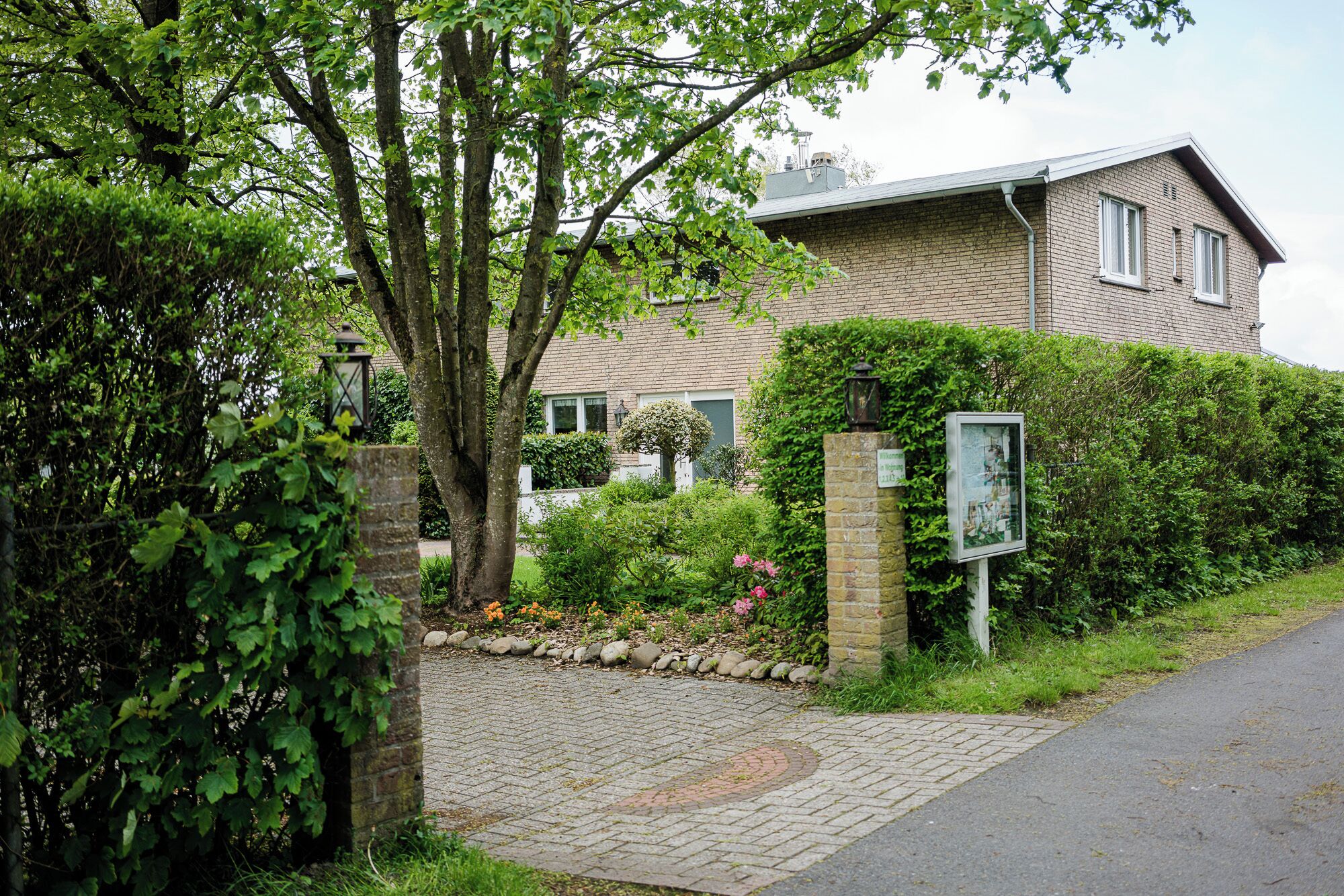 Een woonhuis met een bakstenen gevel, een verharde oprit en een tuin met verschillende planten en bloemen.