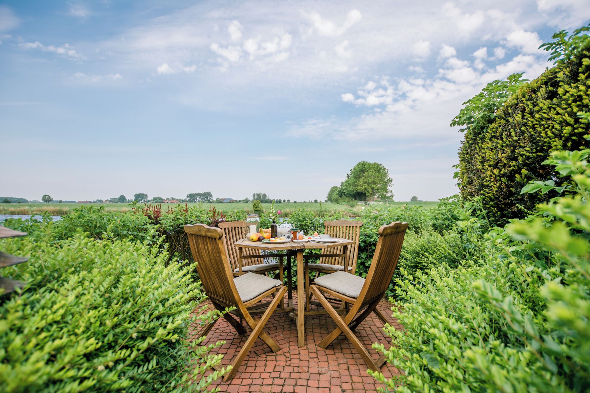 Une table ronde en bois, entourée de chaises, est placée à l’extérieur, au milieu d’une végétation luxuriante, sous un ciel dégagé.