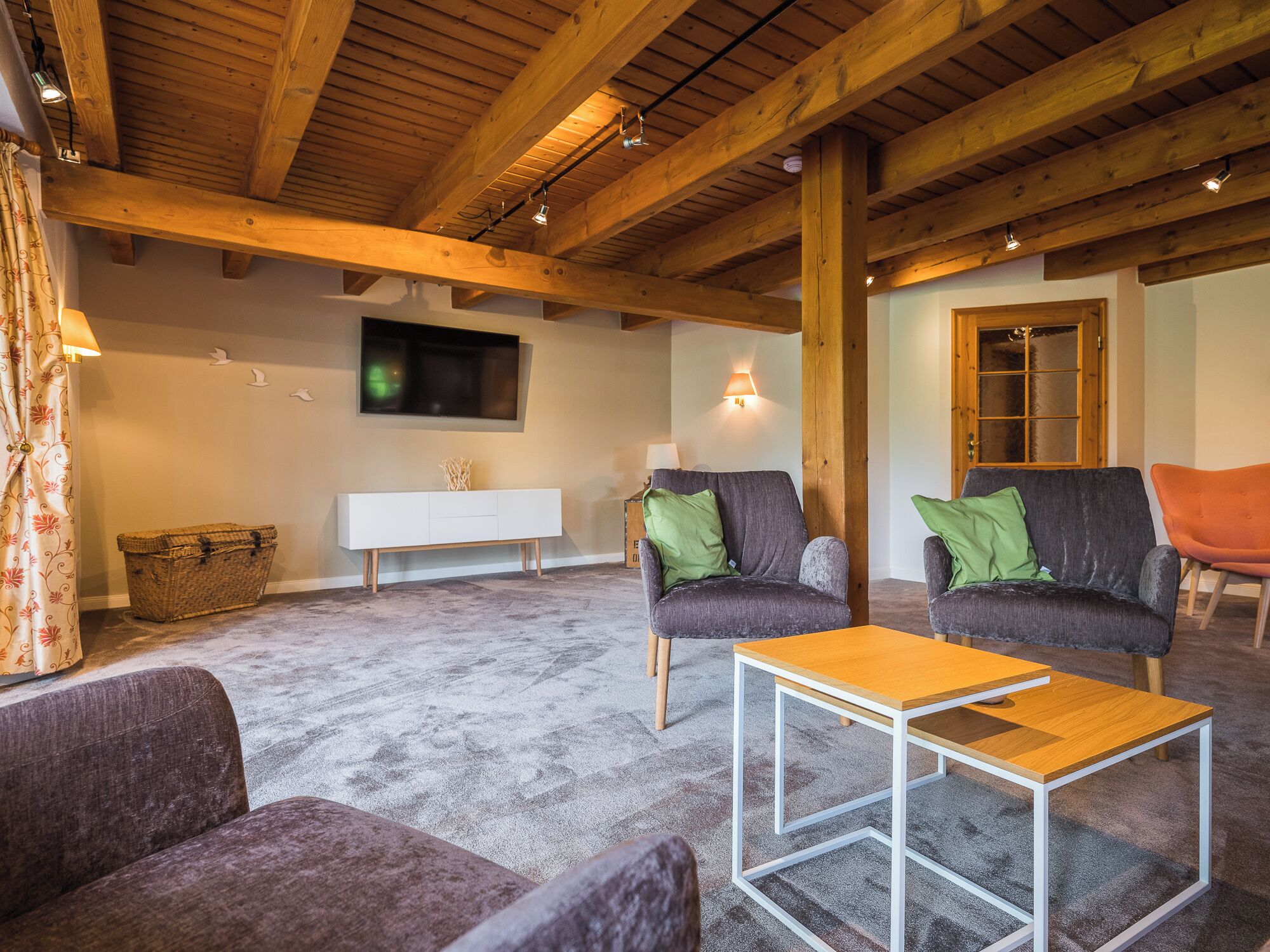 Uma sala de estar espaçosa com teto de madeira alto, um console branco, dois sofás cinzas com almofadas verdes e uma mesa de centro de madeira.