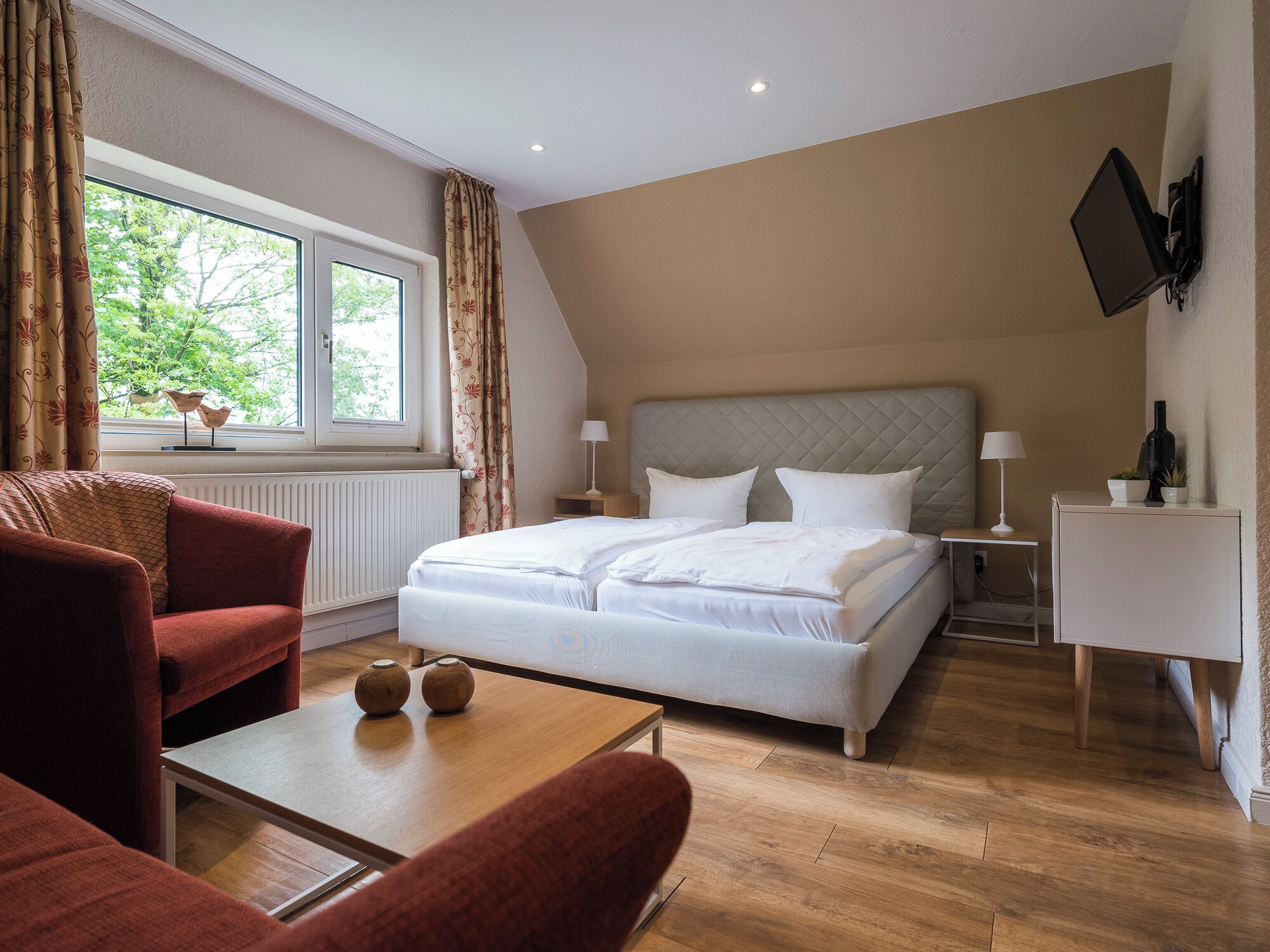 A modern bedroom with a bed, a TV mounted on the wall, a window with curtains, and a wooden floor.