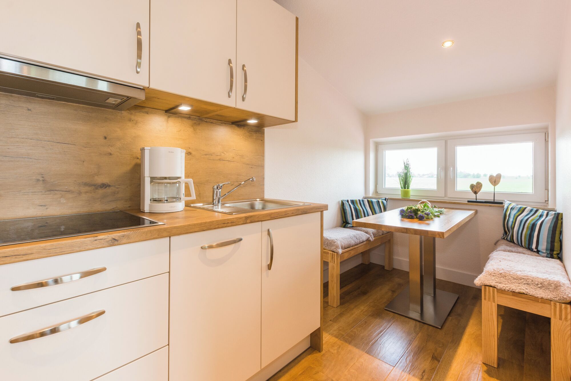 A compact kitchen with a coffee maker, a sink, and wooden cabinets. There is a window with a view of the outdoors and a bench with cushions.