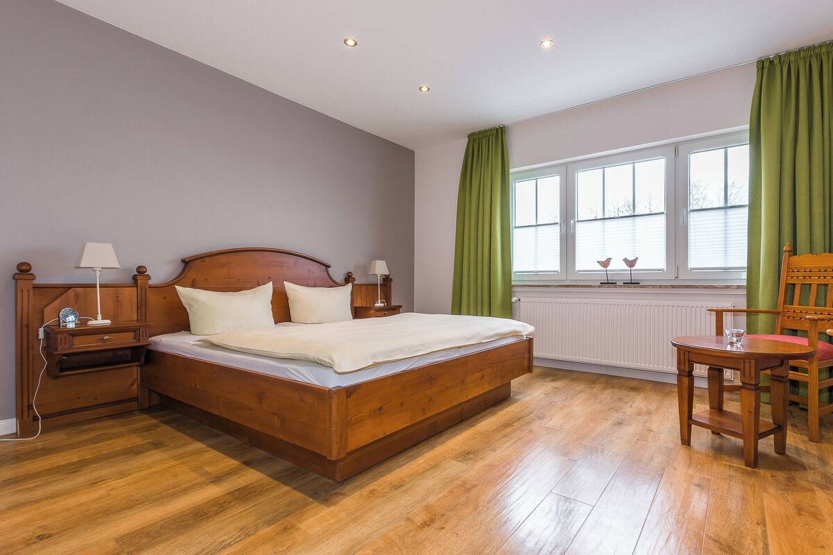 A bedroom with a wooden bed, nightstands, a chair, a window with curtains, and a radiator.