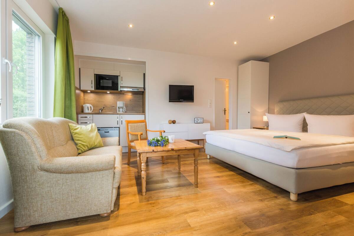 A compact hotel room with a bed, a sofa, a dining table, a kitchenette, and a TV.