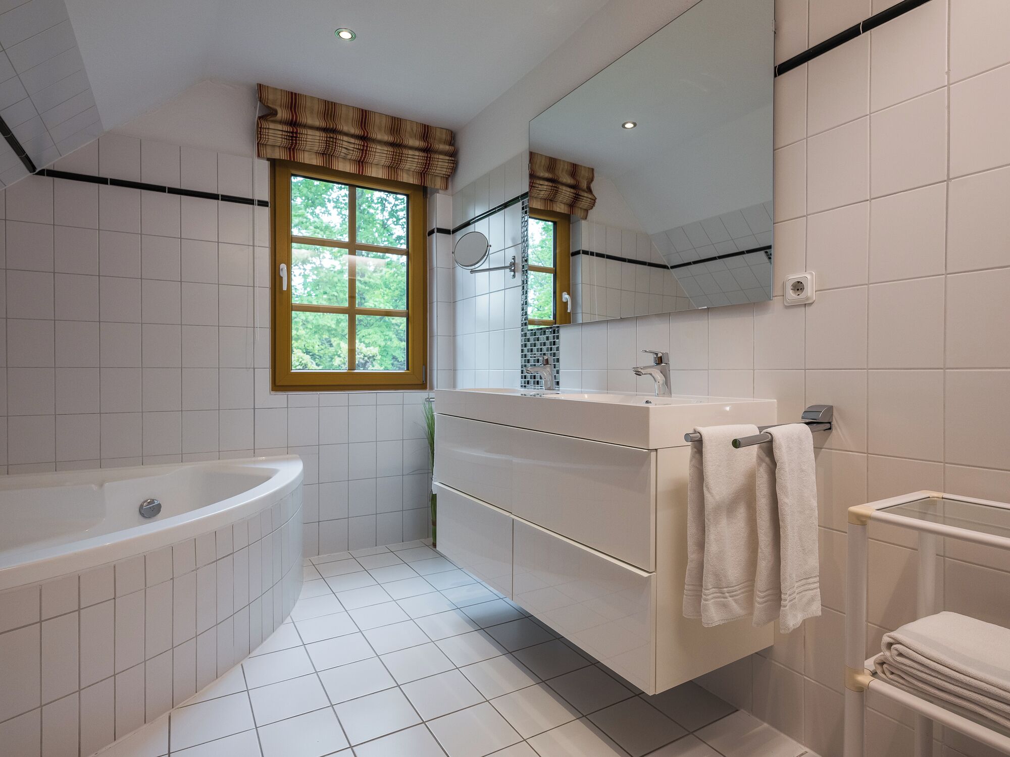 Une salle de bain moderne avec une baignoire, un meuble-lavabo avec un miroir et une fenêtre donnant sur un paysage verdoyant.