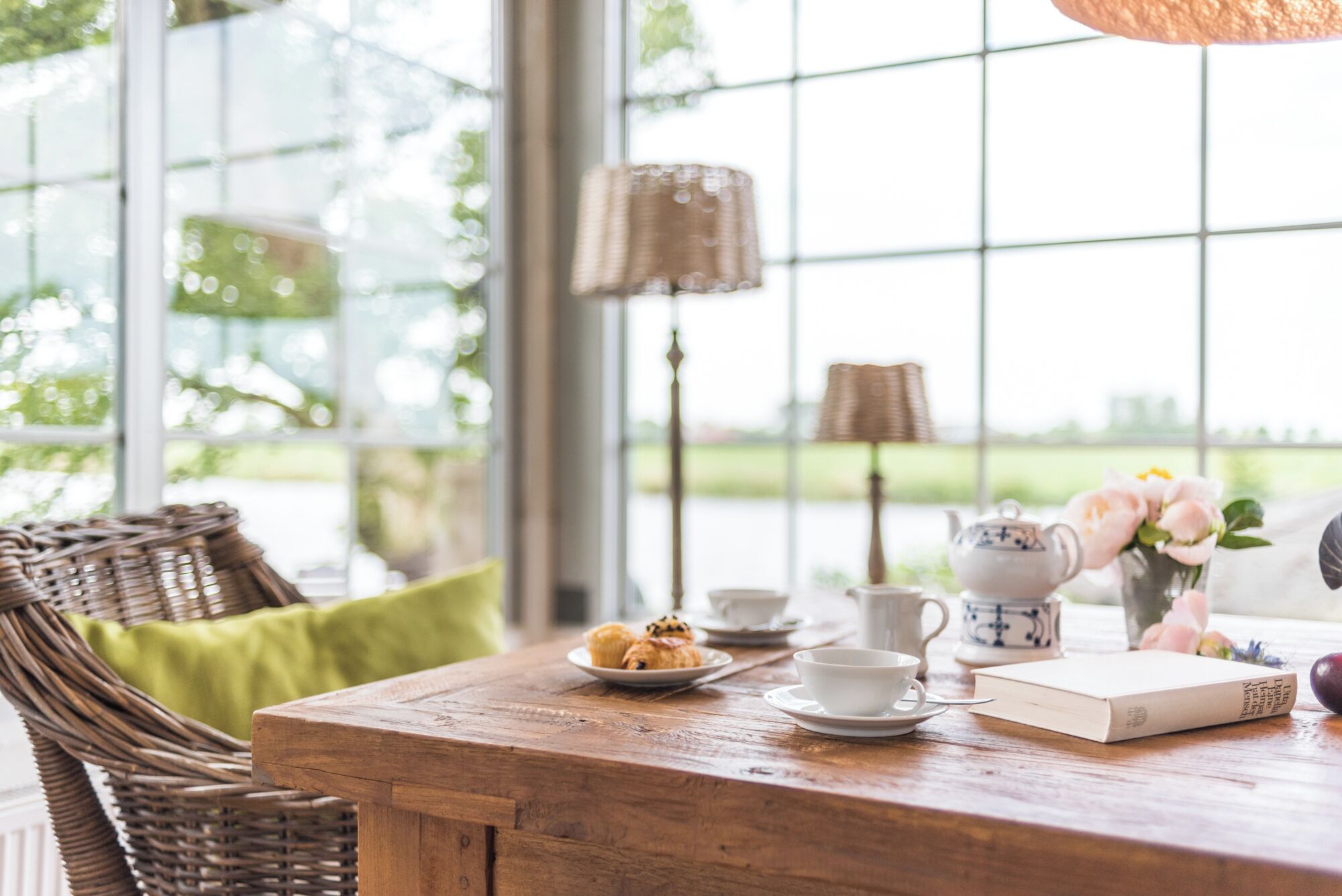 A wooden table with a wicker chair, a green cushion, a teapot, cups, a book, and a plate with pastries.