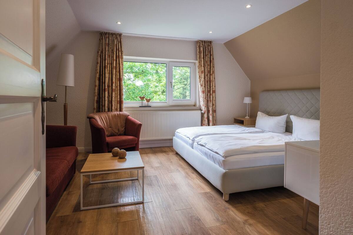 A bedroom with a bed, a chair, a table, a window with curtains, and a radiator.
