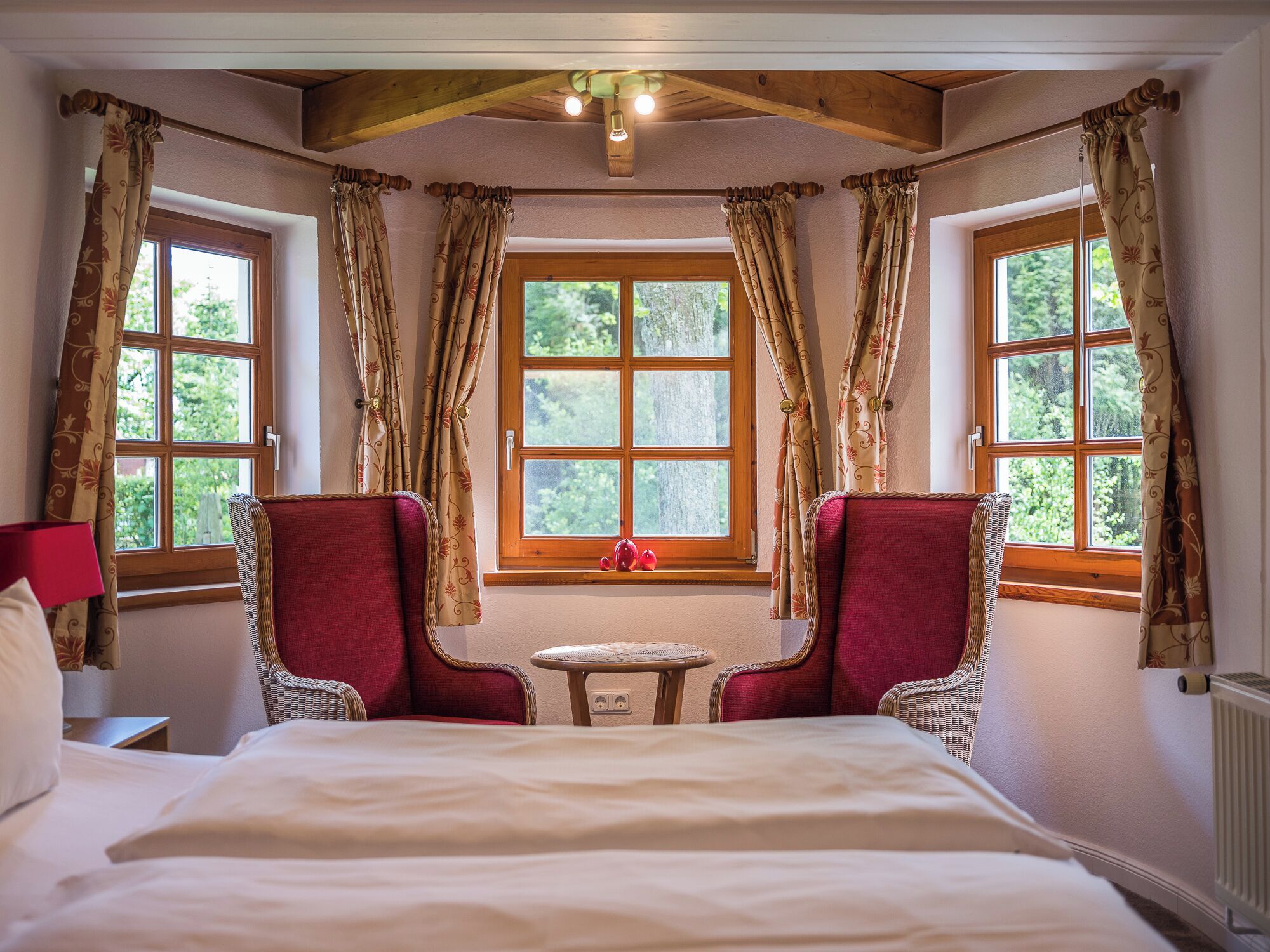 A bedroom with a large bed, two red armchairs, a small table, and two windows with curtains.