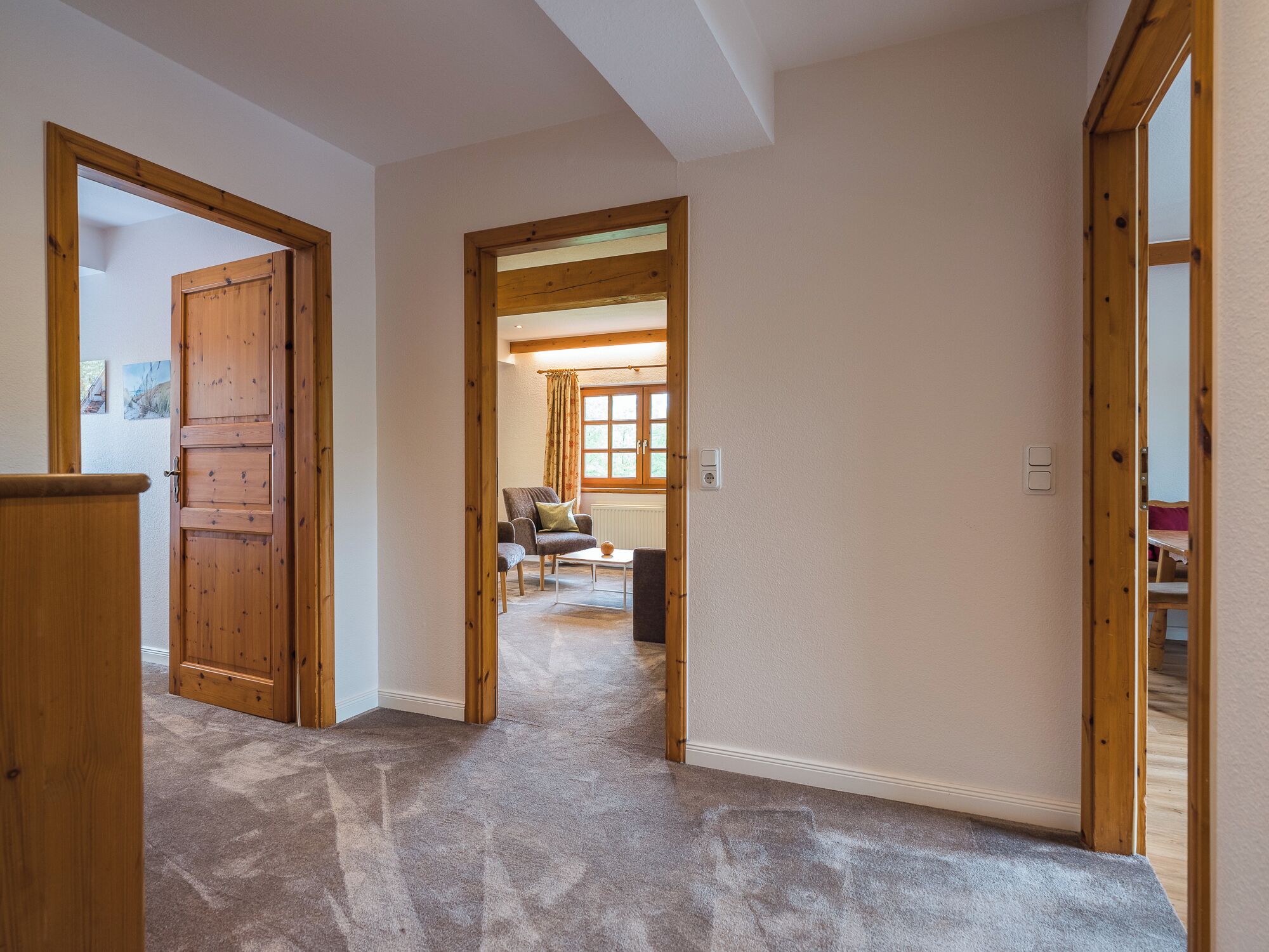 A hallway with wooden doors and a carpeted floor leading to a living area with a chair and a table.