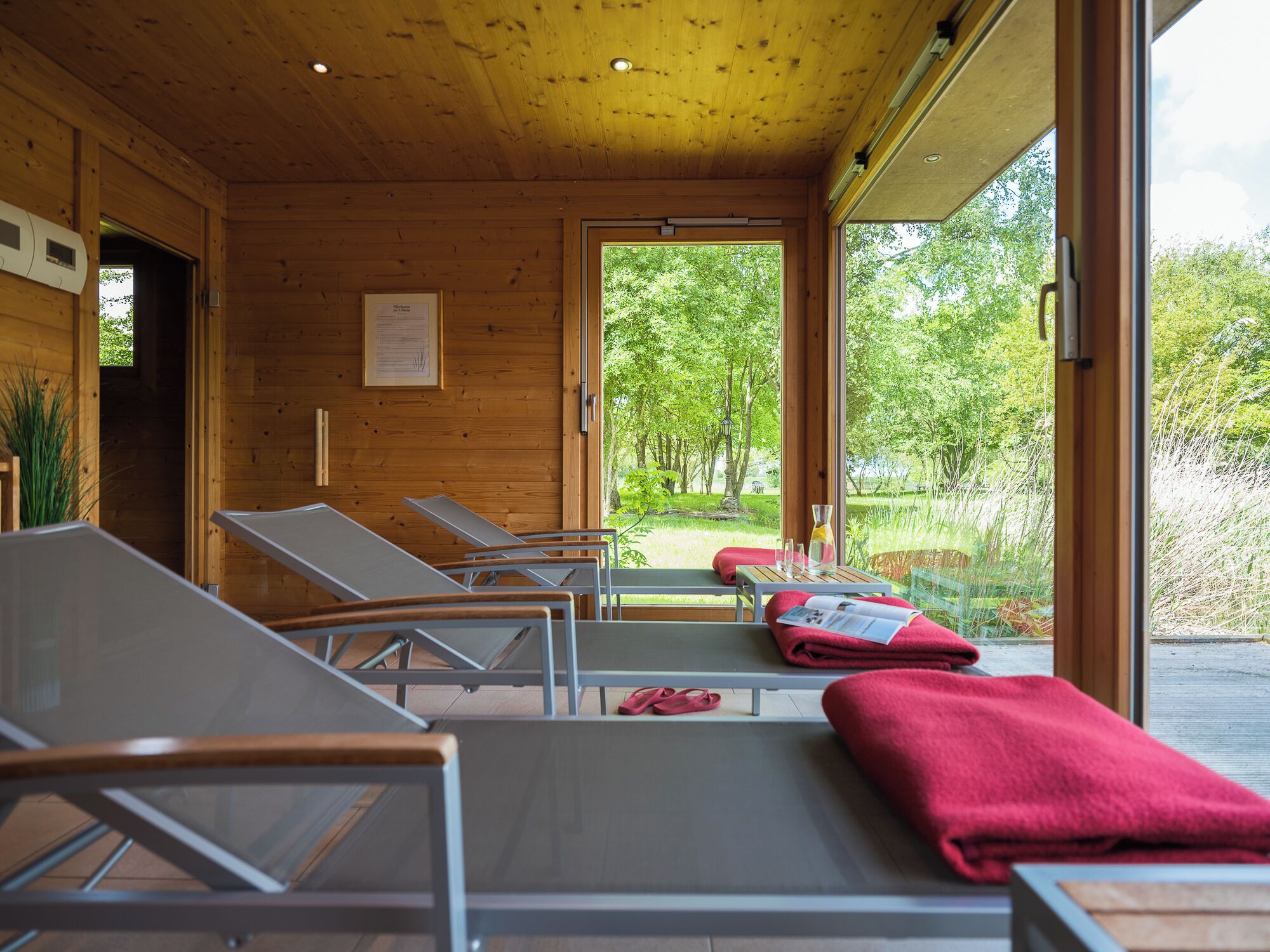 A room with wooden walls and large windows, featuring sun loungers and a view of a green landscape.