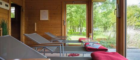 A room with wooden walls and large windows, featuring sun loungers and a view of a green landscape.