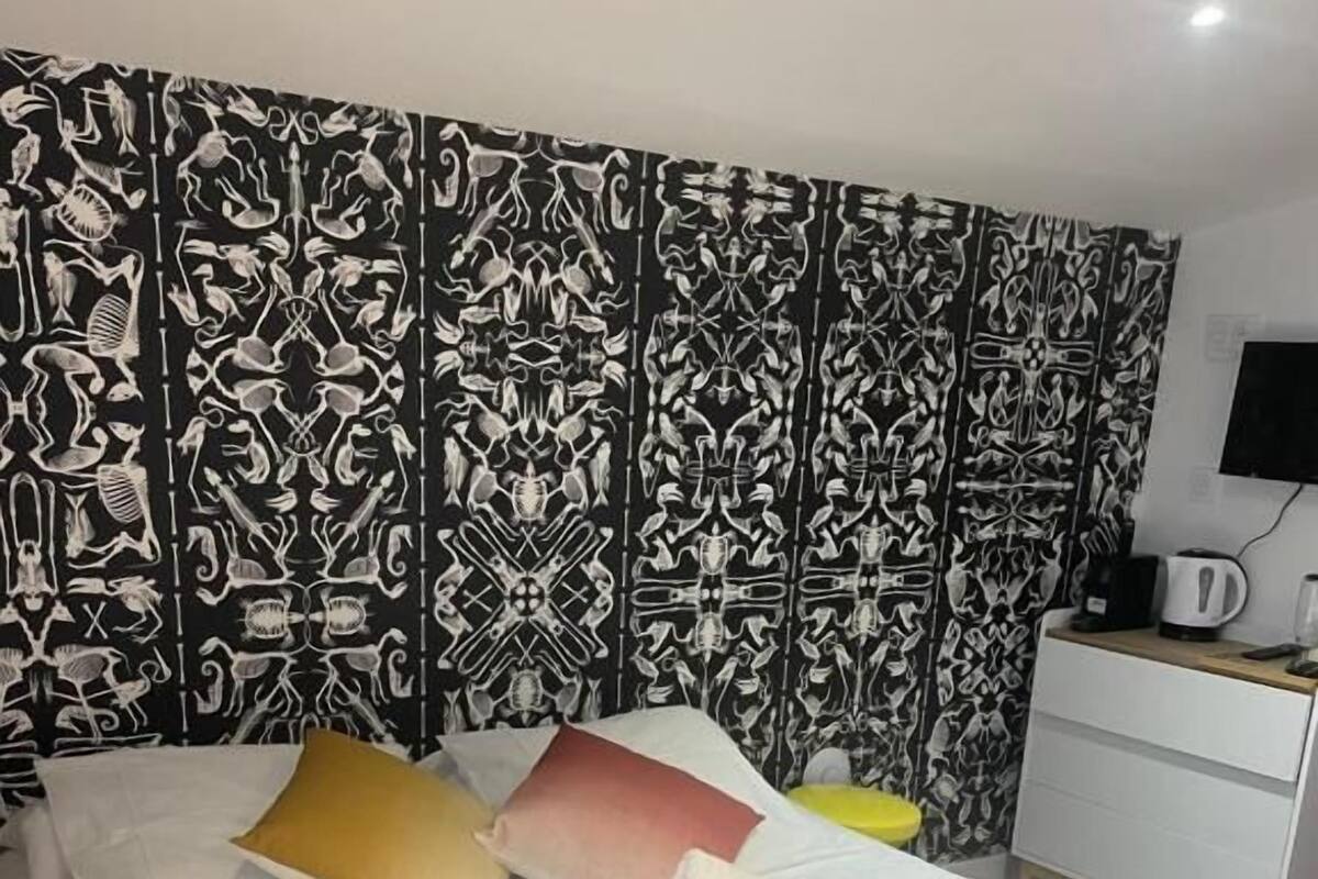 Una habitación de hotel con cama, almohadas, una mesita de noche y una tetera, además de un papel tapiz estampado.