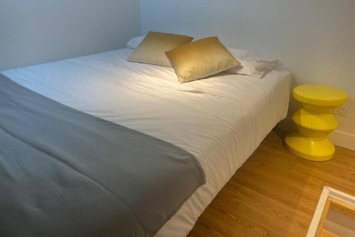 Una cama con ropa blanca, dos almohadas, un plaid gris y una mesita de noche amarilla con una lámpara.