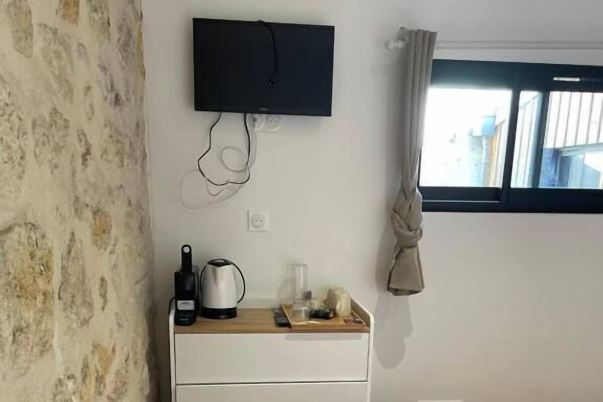 Un dormitorio con cama, mesita de noche, televisor montado en la pared y ventana con cortinas.