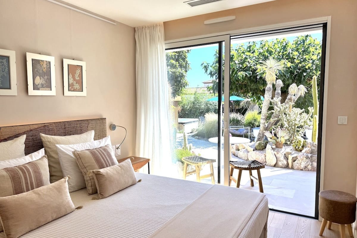 A bedroom with a large bed, a nightstand, a window with a view of a garden, and framed pictures on the wall.