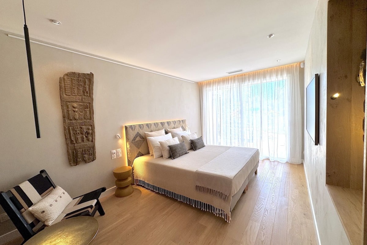 A modern hotel room with a large bed, a wooden floor, a wall-mounted TV, and a decorative headboard.