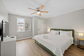 Un dormitorio con cama, ventilador de techo, televisor, escritorio y ventana.