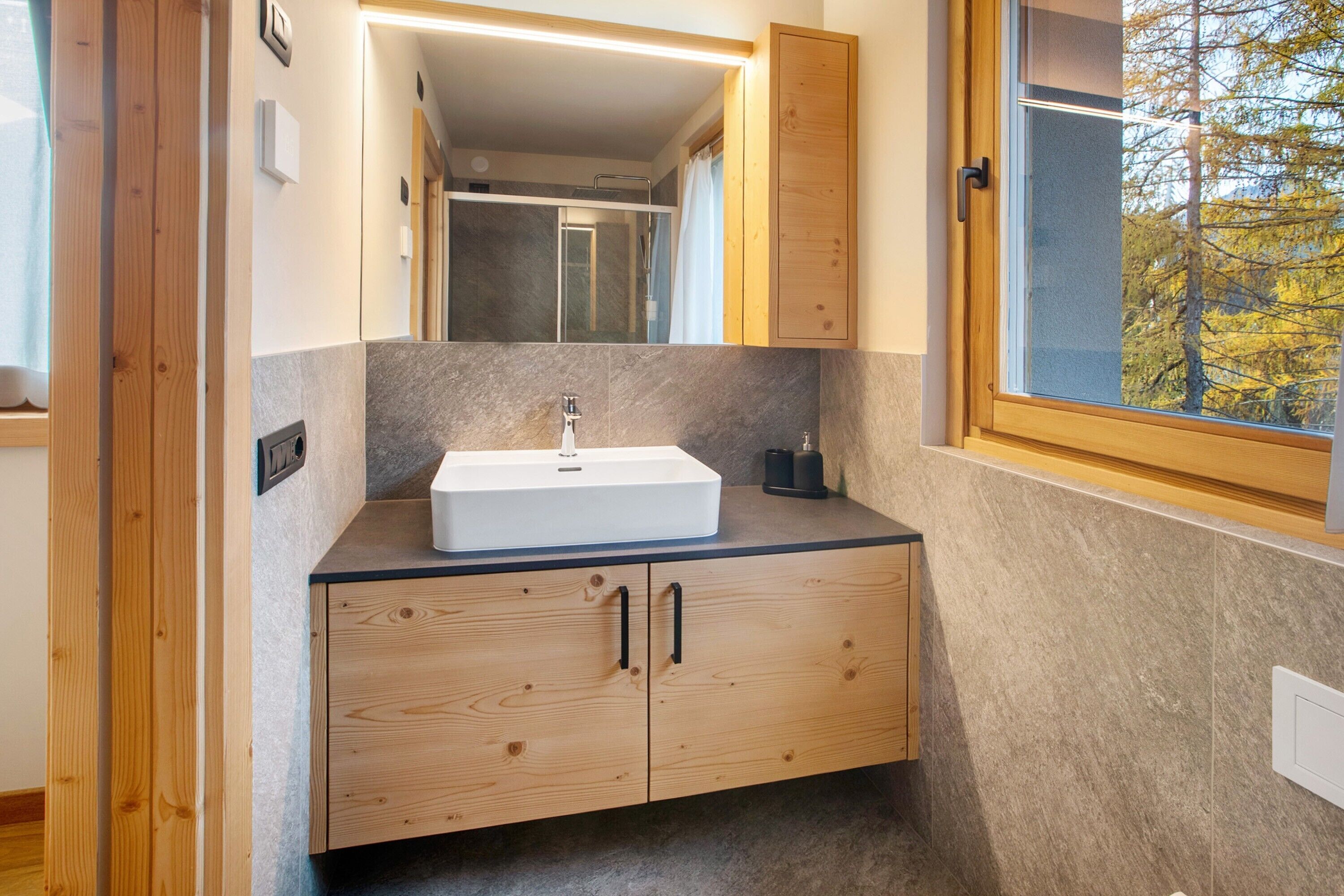 Ein Badezimmer mit einem Holzkosmetikschrank, einem weißen Waschbecken und einem Fenster mit Blick auf Bäume.